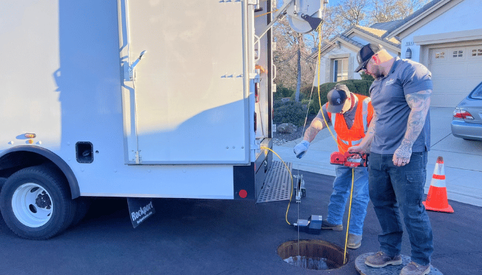 Two workers in high-visibility safety gear stand near a large white truck while one uses a tool beside a circular manhole cover on the asphalt, with traffic cones and a blue car nearby